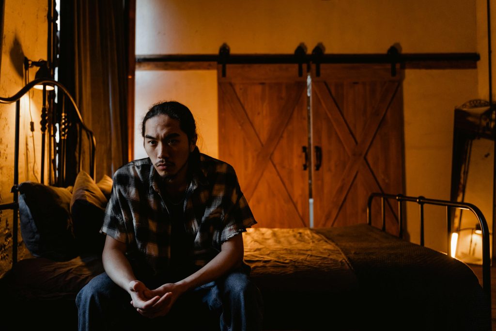 Pensive man sitting on bed in rustic, dimly lit room, creating a reflective mood.